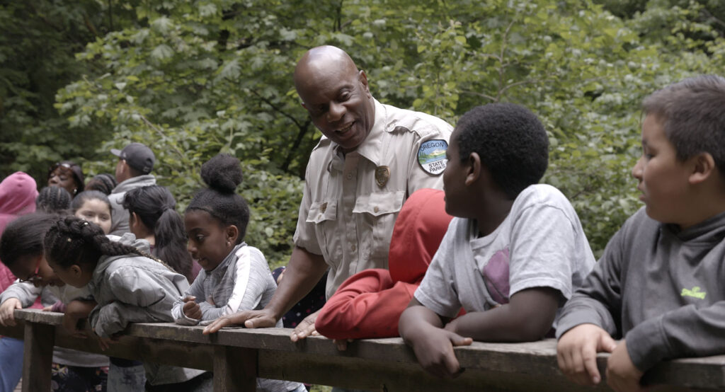 A forest ranger talks to children while standing on a bridge in the forest.