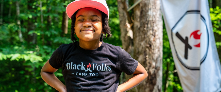A young Black girl with a Black Folks Camp Too shirt on smiles in the forest.